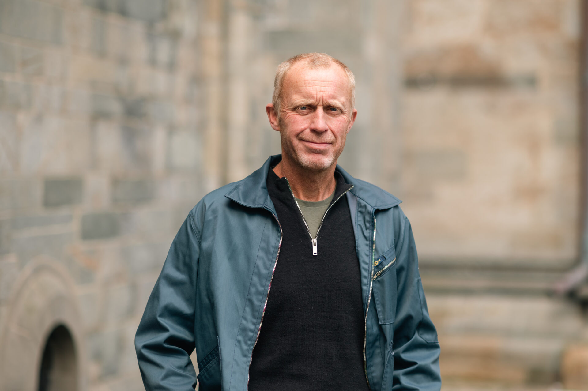 En mann står og smiler. I bakgrunnen ser vi en murvegg.