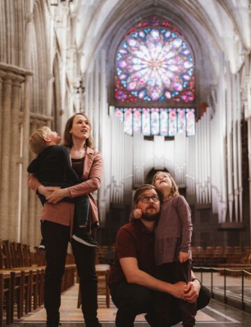 En familie med to små barn står inne i en katedral og ser opp i taket. I bakgrunnen ser vi et stort glassmaleri.