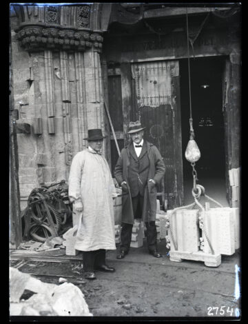 Two men in vintage attire stand outside a damaged stone building with ornate columns; one wears a long coat and bowler hat, the other a suit and fedora. industrial equipment and a hanging scale with a rope are nearby. Two men in vintage attire stand outside a damaged stone building with ornate columns; one wears a long coat and bowler hat, the other a suit and fedora. industrial equipment and a hanging scale with a rope are nearby.