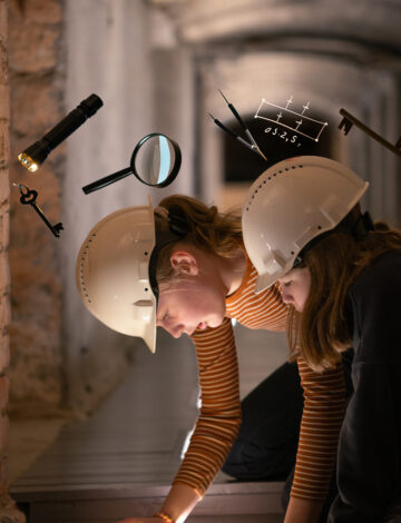 To jenter med hjelm undersøker noe. I bildet ser vi også en lommelykt, en nøkkel og et forstørrelsesglass.