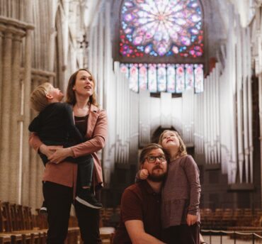 En familie med to små barn står inne i en katedral og ser opp i taket. I bakgrunnen ser vi et stort glassmaleri.