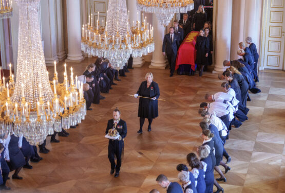 En stor sal med high end seremoni: to rader av kledd personell står bøyd i stille bønn på begge sider av et bredt mønsterbord, mens en kvinne i midten holder en stav og en person foran bærer en innkledd krone. Høye, glitrende lysekroner henger fra taket og edel stil i rommet.
