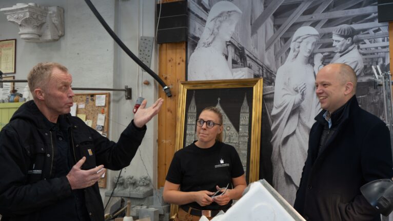 Bilde fra et steinhuggerverksted. En mann forklarer, en kvinnelig steinhugger og tidligere finansminister Trygve Slagsvold Vedum følger med.