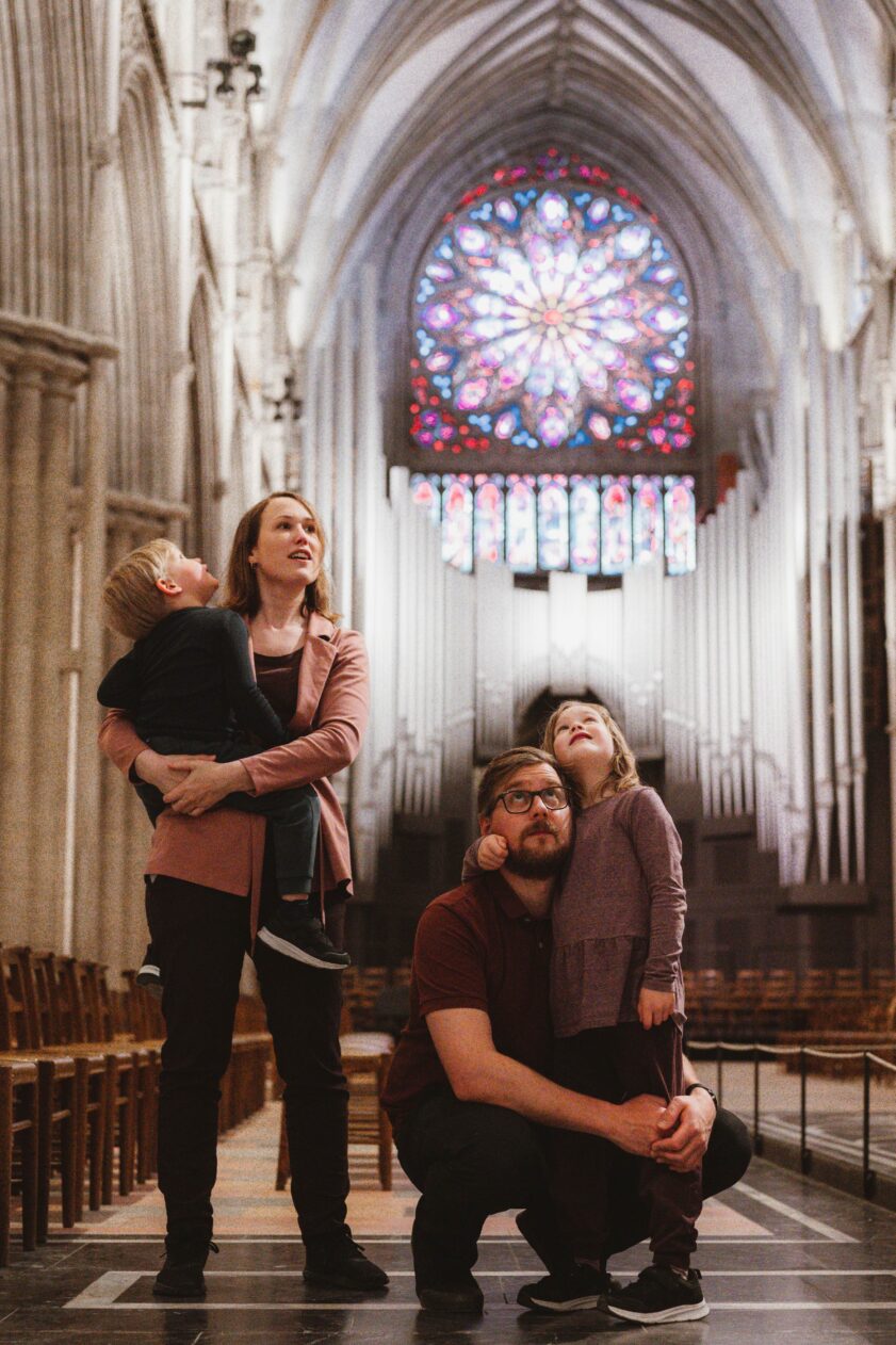 En familie med to små barn står inne i en katedral og ser opp i taket. I bakgrunnen ser vi et stort glassmaleri.