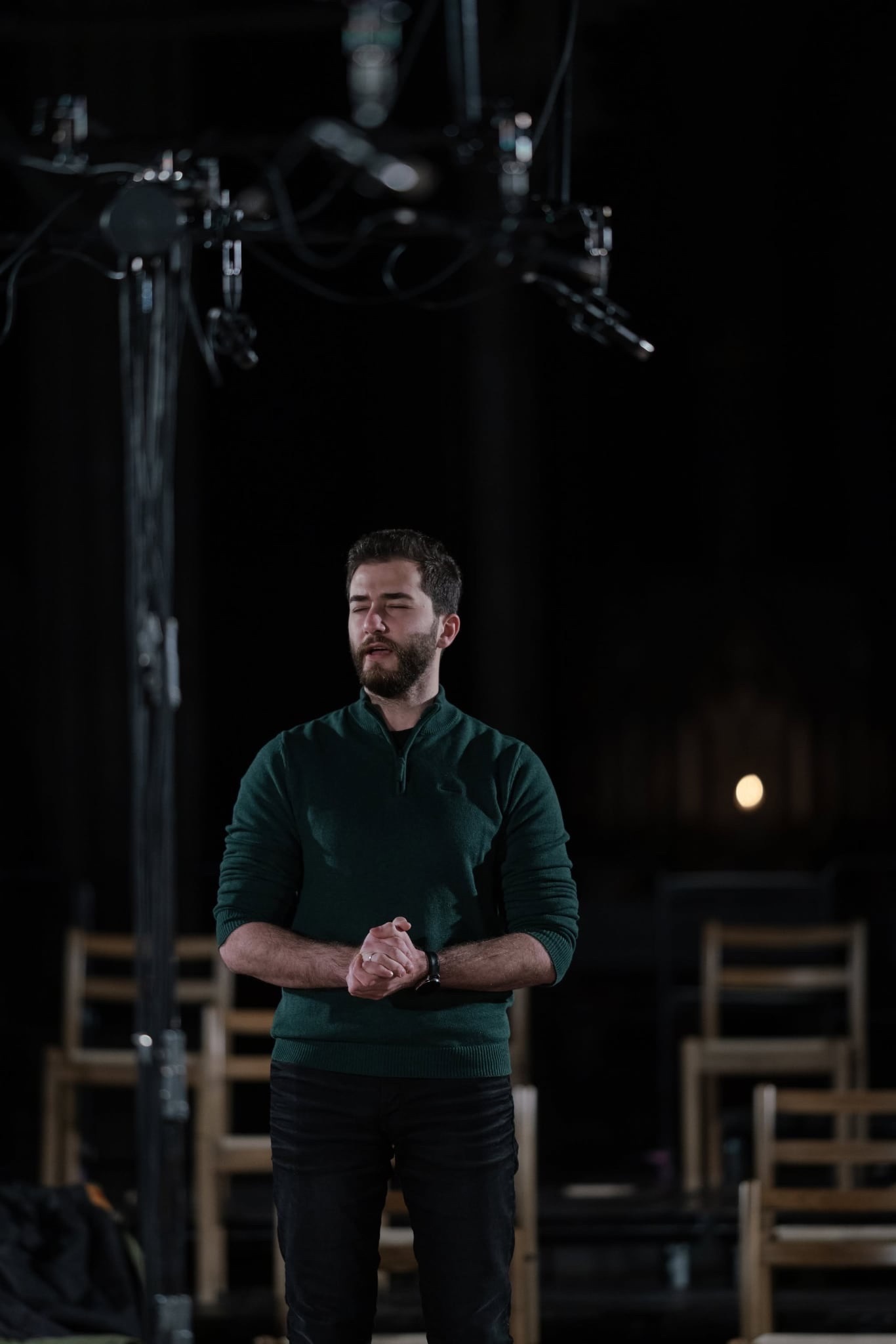 en mann står på scenen i mørket, iført grønn genser og svarte bukser, med lukkede hender og lukket øyne, bak scenetrekk og teaterstoler synlig i bakgrunnen en mann står på scenen i mørket, iført grønn genser og svarte bukser, med lukkede hender og lukket øyne, bak scenetrekk og teaterstoler synlig i bakgrunnen