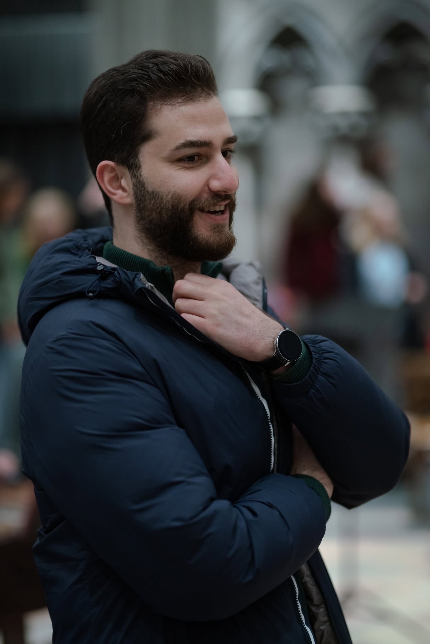 En mann med kort mørkt hår og skjegg smiler mens han justerer den mørkeblå jakken; han står utendørs i en bymiljø. En mann med kort mørkt hår og skjegg smiler mens han justerer den mørkeblå jakken; han står utendørs i en bymiljø.