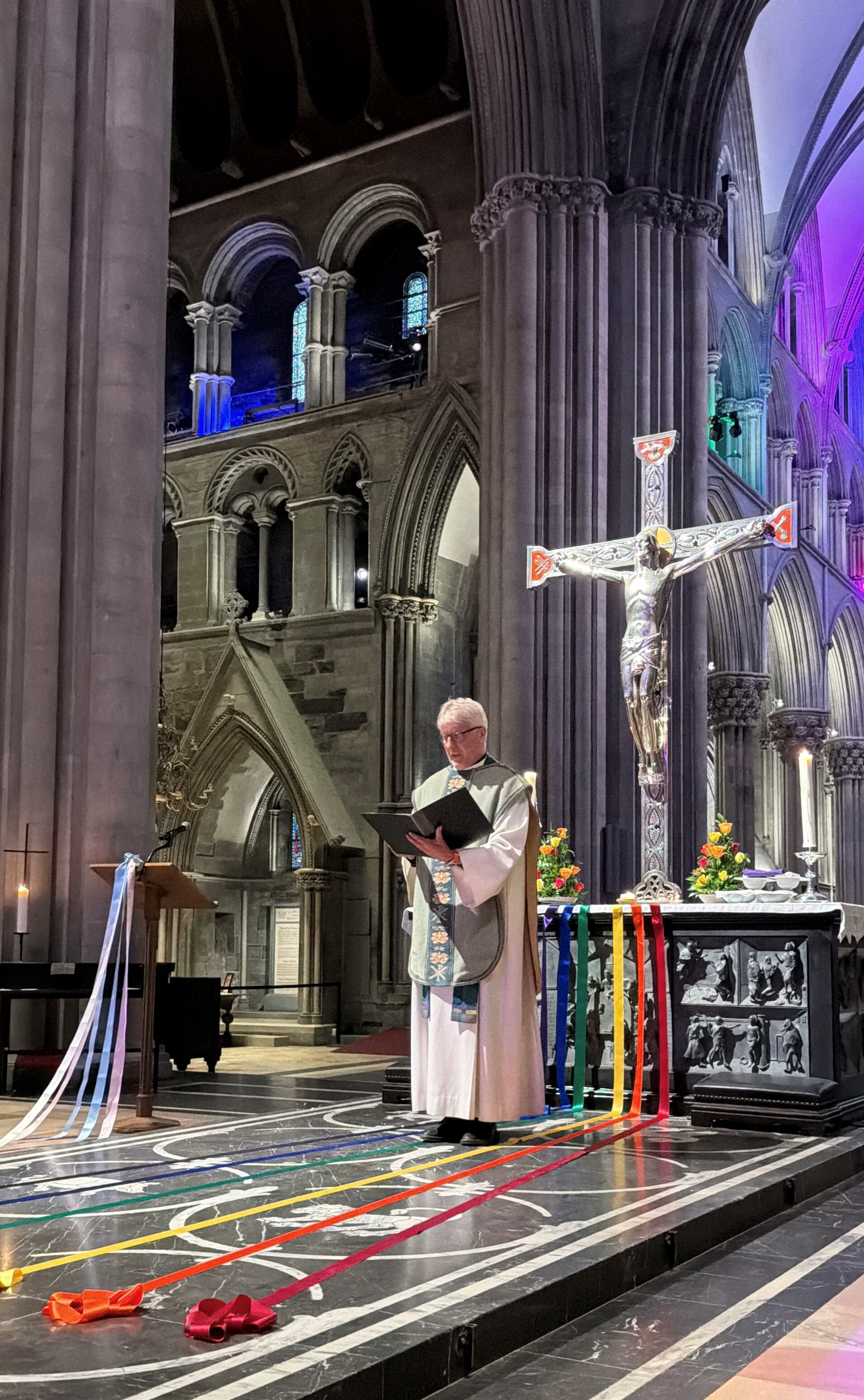 Kledd i liturgisk drakt står en prest ved alteret i en gotisk katedral og leser fra en bok, omgitt av fargerike bånd og et stort krusifiks.