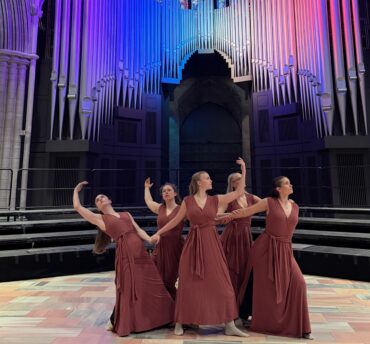 Fem dansere i lange brune kjoler står på en stort åpnet scene i en katedrallignende hall med et stort pipeorgel i bakgrunnen, farget blått og lilla lys. Fem dansere i lange brune kjoler står på en stort åpnet scene i en katedrallignende hall med et stort pipeorgel i bakgrunnen, farget blått og lilla lys.