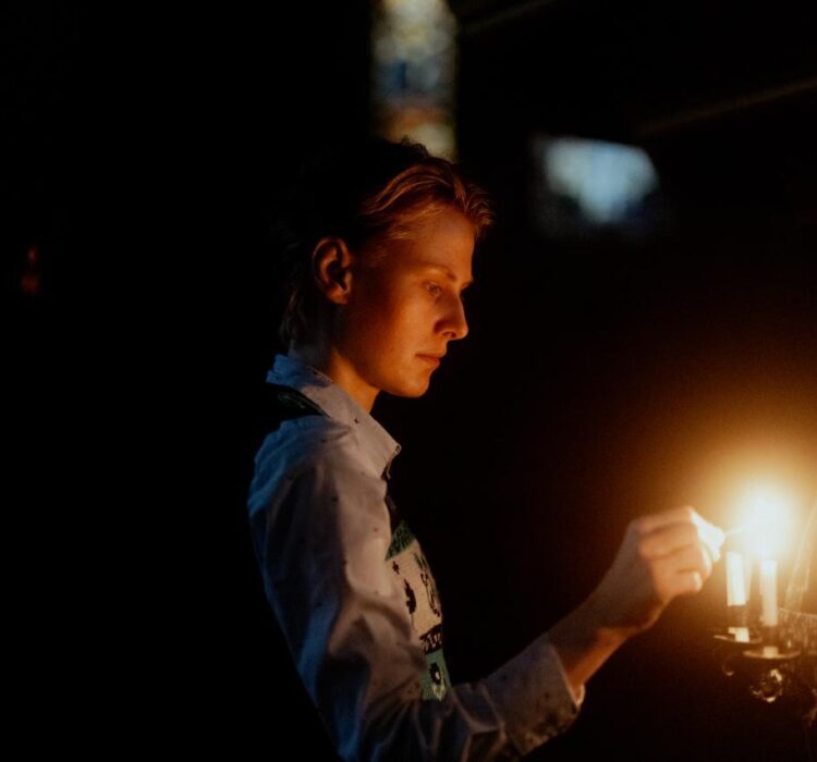 En person står i mørket og tenner lys i en lysekrone, ansiktet opplyst av flammen mens håret gløder i nattlys. En person står i mørket og tenner lys i en lysekrone, ansiktet opplyst av flammen mens håret gløder i nattlys.