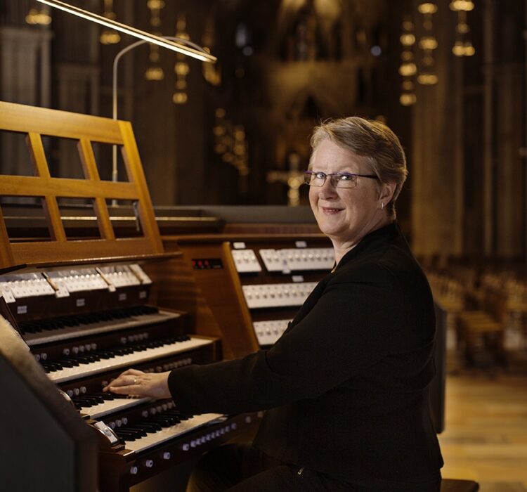 Kvinne spiller på et stort trekorgan i en kirke eller konsertsal, smiling mot kamera, iført svart jakke, med fokusert arbeid ved tangentene.