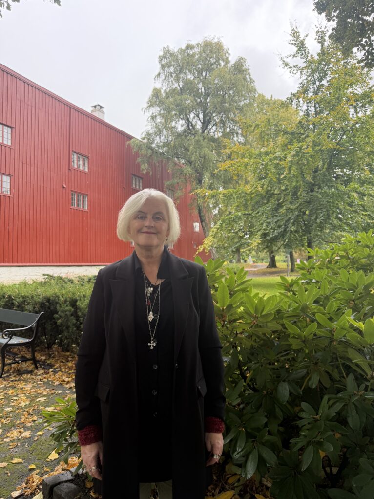 En kvinne med kort lyst hår står ute i hagen ved en rød bygning; hun har svart lang frakk og flere halskjeder, og det ligger gule blader på bakken med grønne busker i forgrunnen.