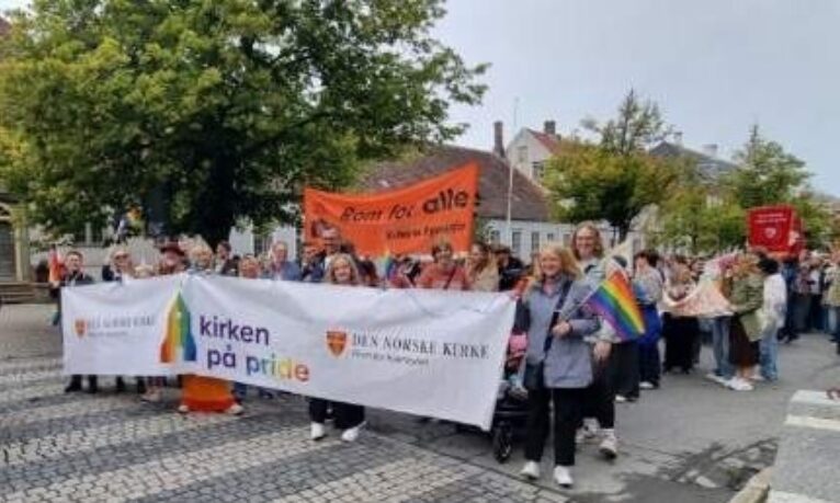gruppe mennesker i gateparade som bærer banner og regnbueflagg, gater, trær og bylige bygninger i bakgrunnen, deltakere rolig samlet og fotografert utendørs