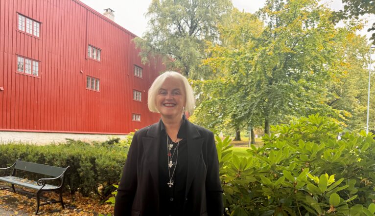En kvinne med kort hvitt hår smiler foran en rød trebygning og grønne busker i en park, iført svart blazer og flerstrengede pe_nny, midt i et grønt landskap. En kvinne med kort hvitt hår smiler foran en rød trebygning og grønne busker i en park, iført svart blazer og flerstrengede pe_nny, midt i et grønt landskap.