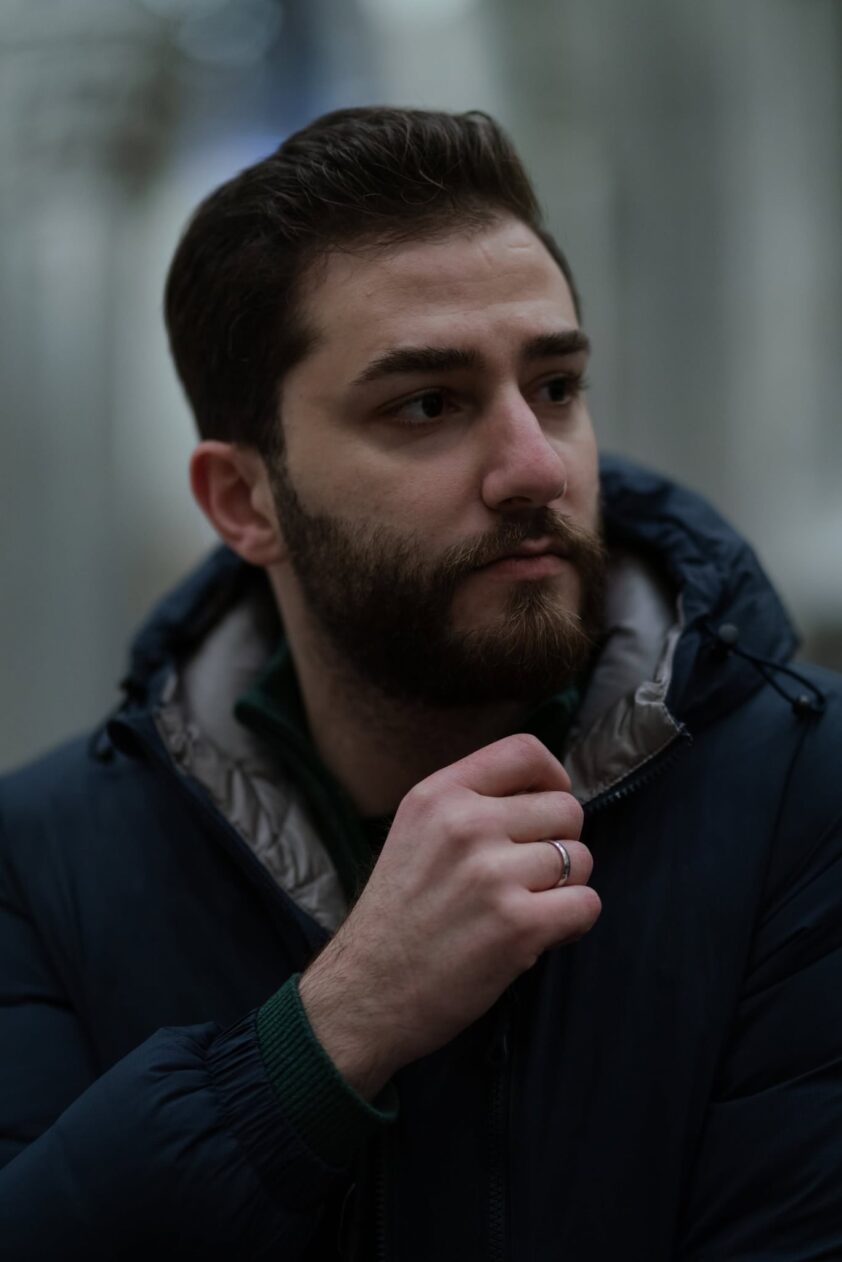 En mann med kort mørkt hår og skjegg, ser til venstre, bærer en mørk jakke og har hendene nær halsen, i utendørs, urban bakgrunn. En mann med kort mørkt hår og skjegg, ser til venstre, bærer en mørk jakke og har hendene nær halsen, i utendørs, urban bakgrunn.