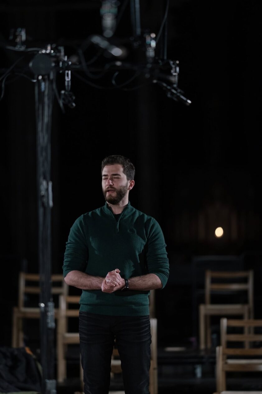 en mann står på scenen i mørket, iført grønn genser og svarte bukser, med lukkede hender og lukket øyne, bak scenetrekk og teaterstoler synlig i bakgrunnen en mann står på scenen i mørket, iført grønn genser og svarte bukser, med lukkede hender og lukket øyne, bak scenetrekk og teaterstoler synlig i bakgrunnen