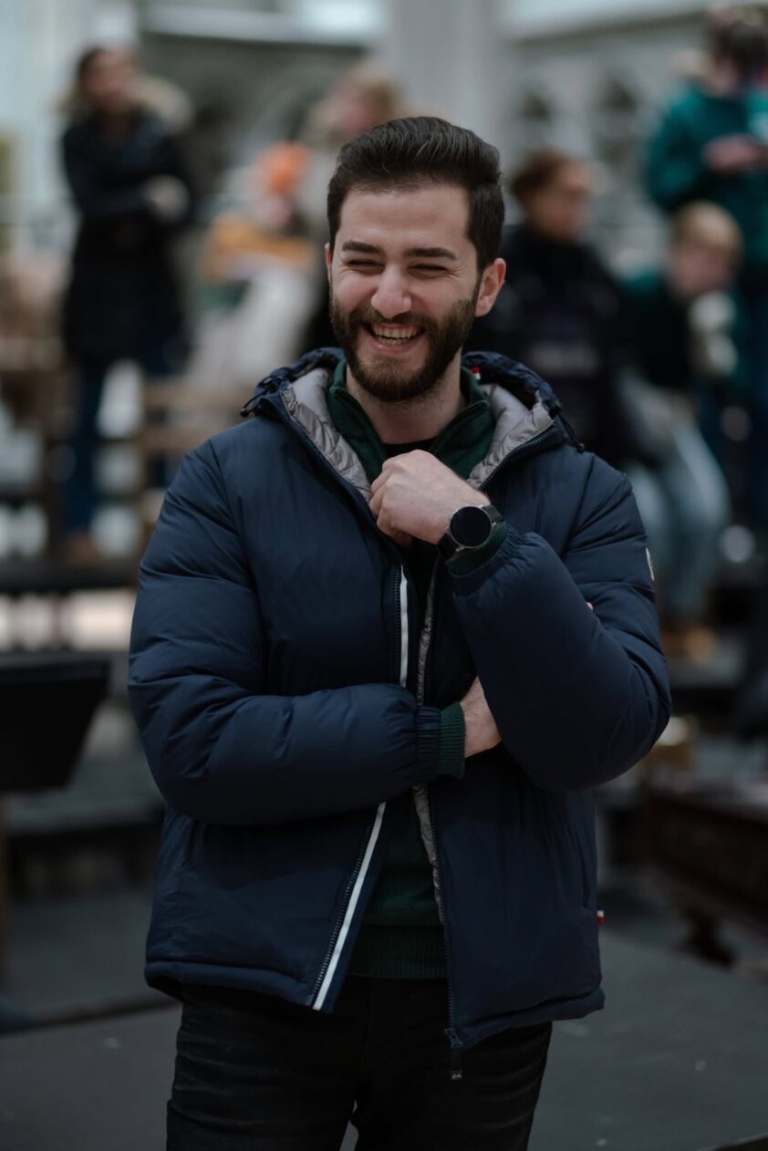 Smilende person i blå jakkke står i en travel butikk eller offentlige plass, hendene holdt nær krage, med tåkelagt bakgrunn og andre mennesker i bakgrunnen. Smilende person i blå jakkke står i en travel butikk eller offentlige plass, hendene holdt nær krage, med tåkelagt bakgrunn og andre mennesker i bakgrunnen.