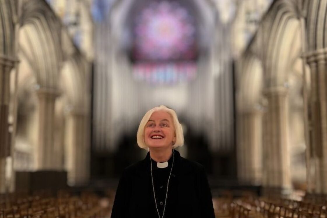 En person med kort lyst hår står inne i en stor gotisk katedral, smiler mens de ser oppover mot en fargerik rosevindu i bakgrunnen.